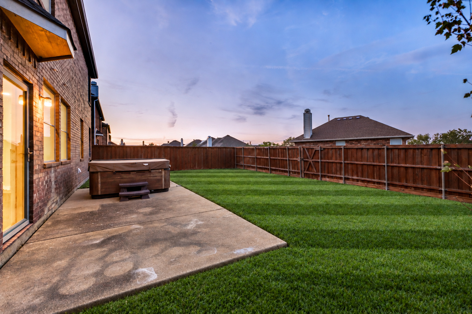Backyard patio of 4215 Maidstone Drive at dusk, showing the covered patio, hot tub, striped lawn and wooden fence.